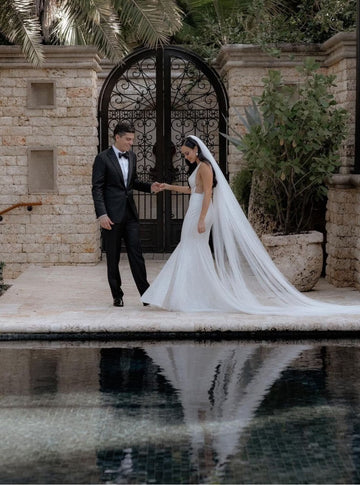Full-length portrait of a bride wearing the sheer, soft Annabelle English net and a fitted mermaid wedding dress. The bride holds the groom's hand as they stand in an outdoor courtyard, with the long, elegant veil reflecting in the water of a pool in the foreground.
