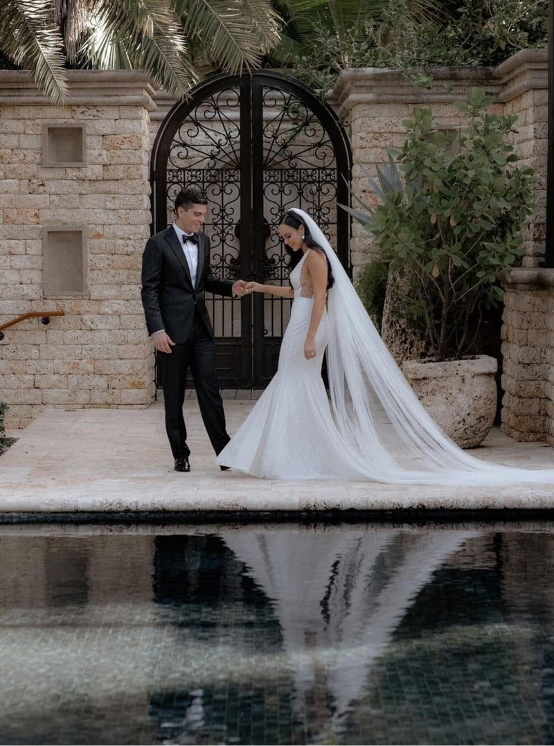 Full-length portrait of a bride wearing the sheer, soft Annabelle English net and a fitted mermaid wedding dress. The bride holds the groom's hand as they stand in an outdoor courtyard, with the long, elegant veil reflecting in the water of a pool in the foreground.