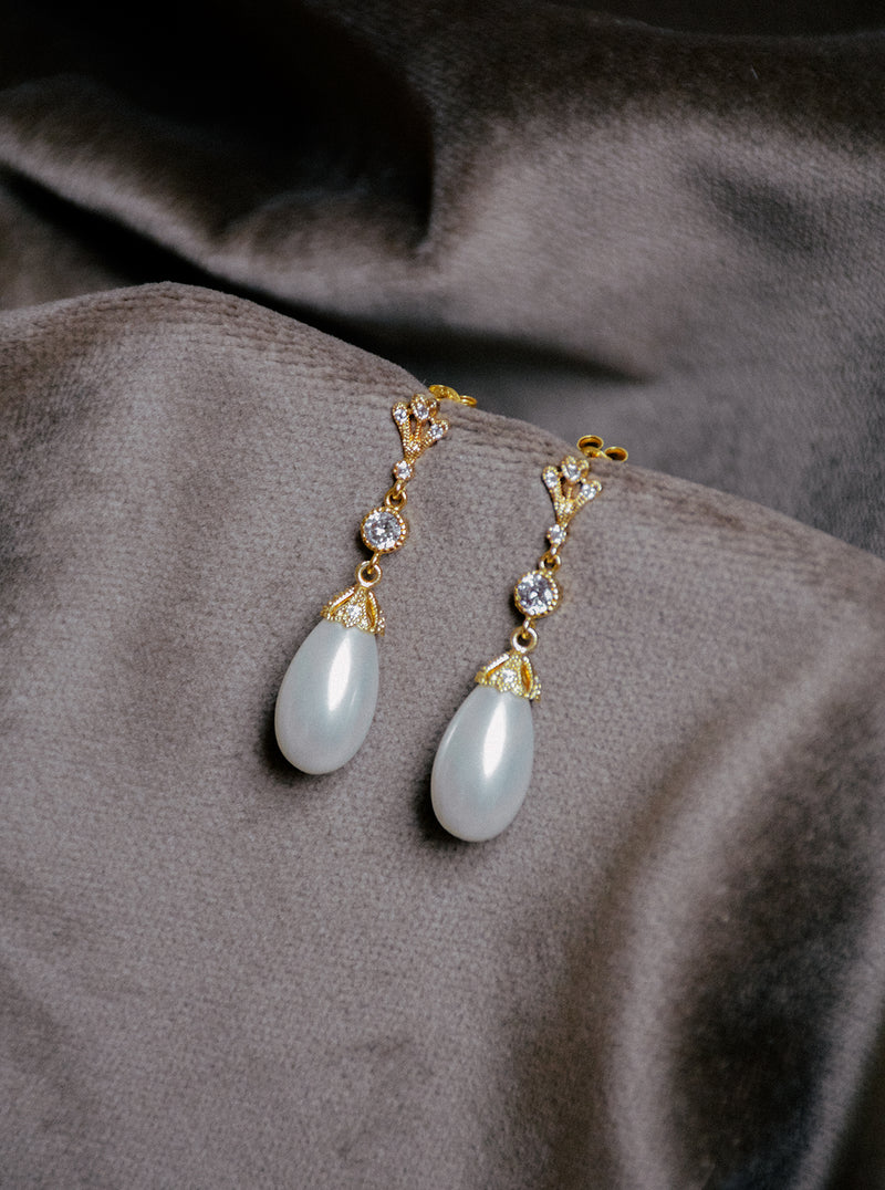 A pair of LYON bridal drop earrings featuring a gold setting, clear crystals, and an oval teardrop pearl resting on textured velvet fabric.