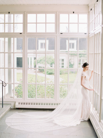 A bride in a lace gown standing in the sunroom at Langdon Hall, with the Olivia long wedding veil fanned out dramatically on the slate floor behind her.