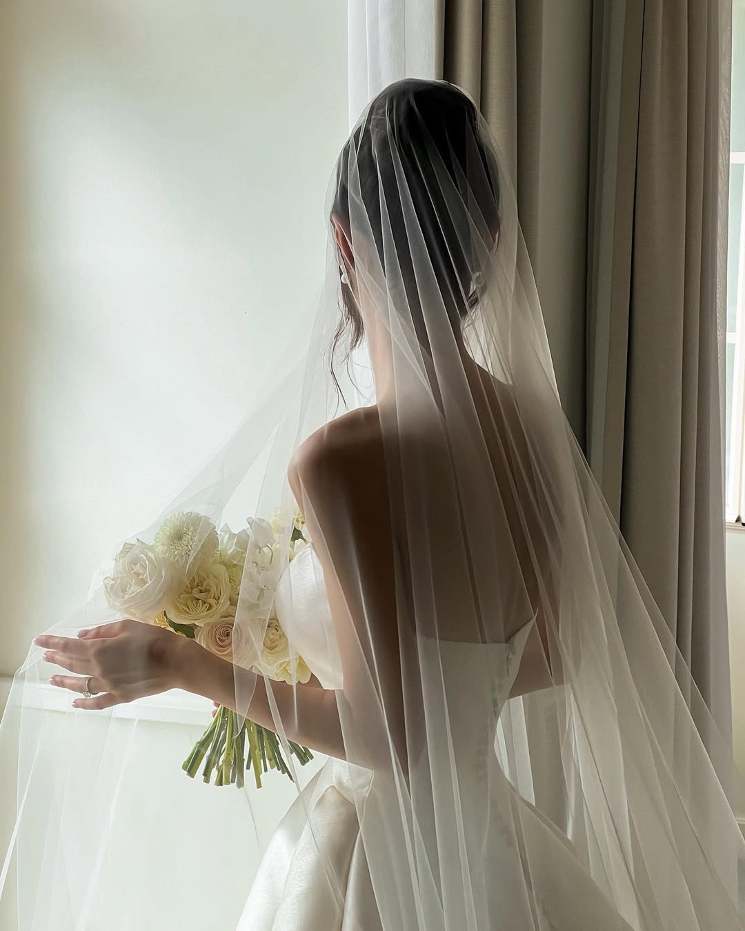 A bride wearing a long wedding veil and a strapless ballgown dress is standing in a softly lit room. She is holding a bouquet of peonies.