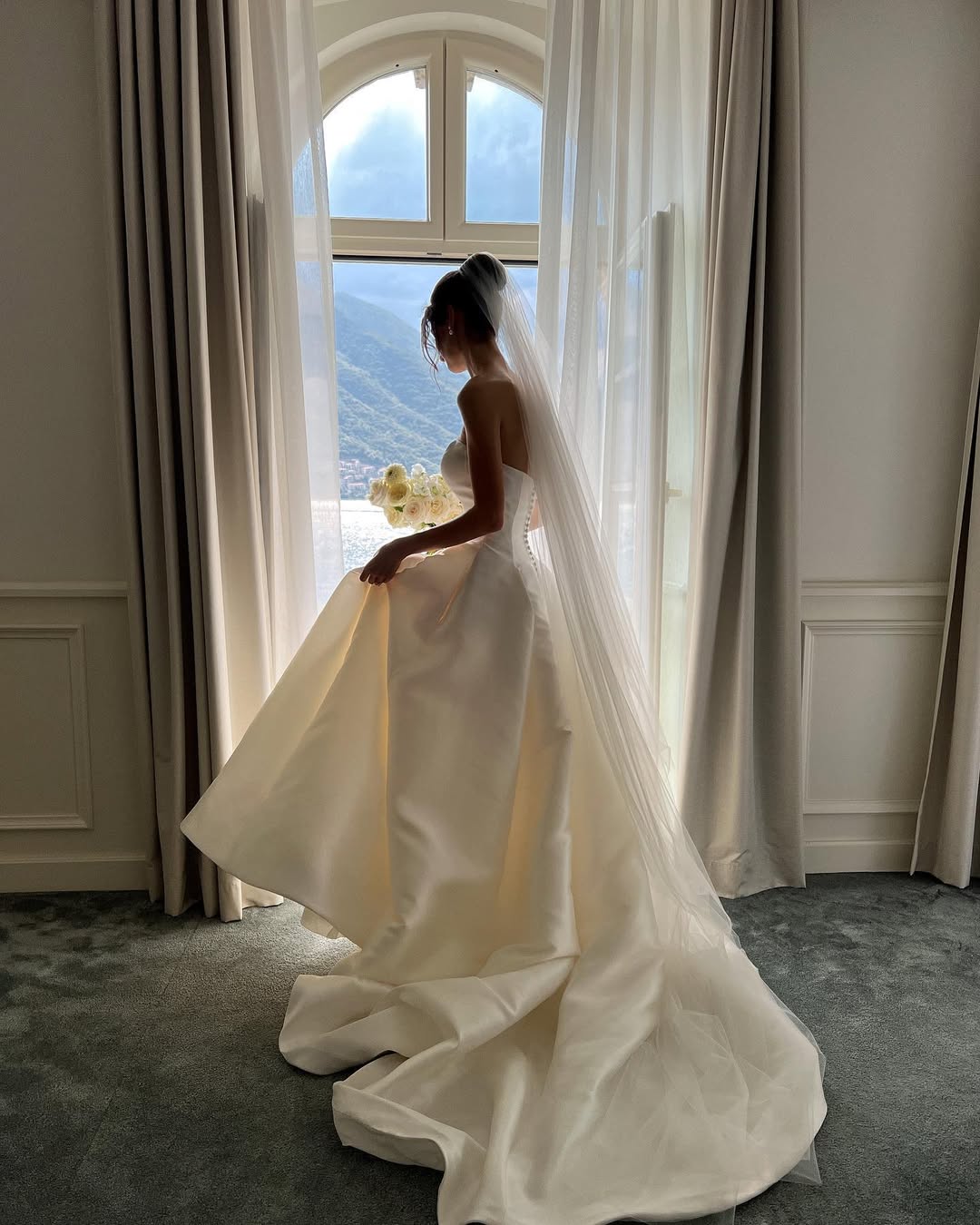 A bride wearing a long wedding veil and a strapless ballgown dress is standing is standing by a tall window with a scenic view.