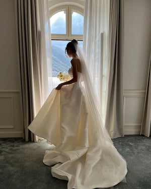 A bride wearing a long wedding veil and a strapless ballgown dress is standing is standing by a tall window with a scenic view.