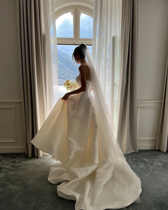 A bride wearing a long wedding veil and a strapless ballgown dress is standing is standing by a tall window with a scenic view.