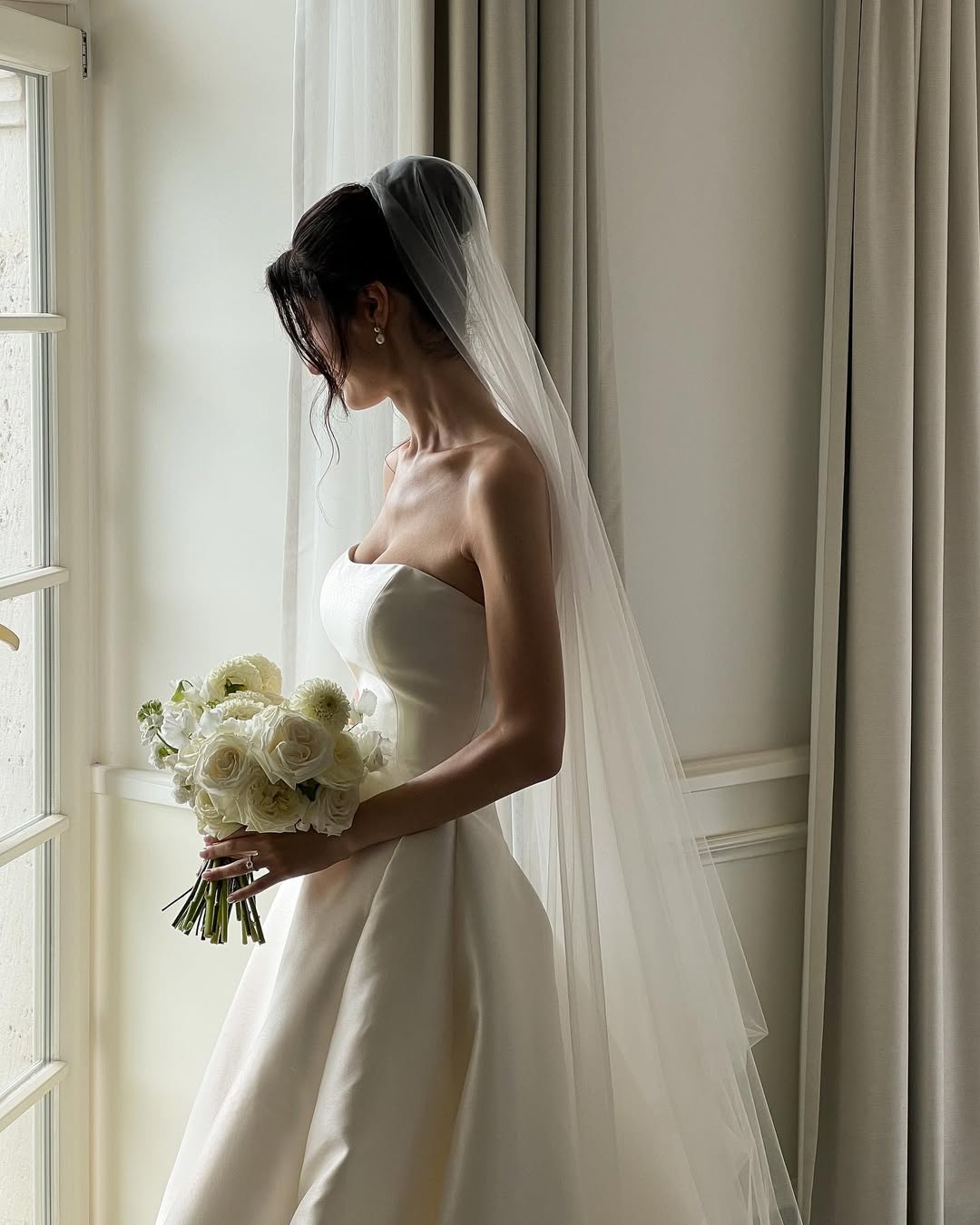 A bride wearing a long wedding veil and a strapless ballgown dress is standing next to a sunny window.