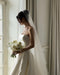 A bride wearing a long wedding veil and a strapless ballgown dress is standing next to a sunny window.