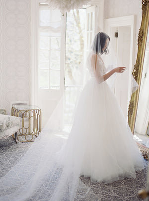 Full-length shot in Graydon Hall Manor of a bride looking into a large gold mirror. The bride is wearing the AMELIE Bridal Veil with Blusher and a tulle ballgown. 