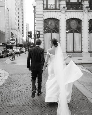 Black and white photo of a real bride and groom walking hand-in-hand on a city street. The long AMELIE Veil with Blusher flows out dramatically behind the bride.