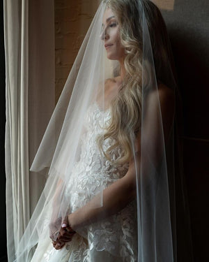 Close-up of a blonde bride with long hair standing by a window. She's wearing the AMELIE bridal veil over her face.