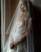 Close-up of a blonde bride with long hair standing by a window. She's wearing the AMELIE bridal veil over her face.
