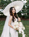 Smiling bride outdoors, holding a white parasol and a bouquet of roses and orchids. She wears the AMELIE Bridal Veil with a lace fit-and-flare gown.