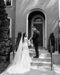 Black and white photo of the bride and groom standing on the steps of The Cordelle. The long AMELIE Veil with Blusher trails behind the bride.