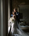 Romantic portrait of the bride and groom embracing by a large window indoors. The bride holds a cascading bouquet and wears the AMELIE Veil with blusher and a lace mermaid gown.