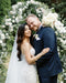 Close-up portrait of the bride and groom embracing in front of a white floral arch. The bride wears the AMELIE Bridal Veil with a strapless lace gown and hair in soft waves.