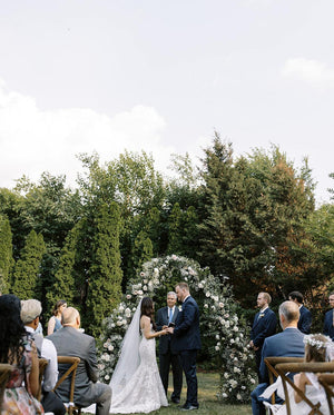 Bride and groom standing beneath a white floral arch during an outdoor ceremony. The sheer AMELIE Veil falls gracefully down the bride's back.