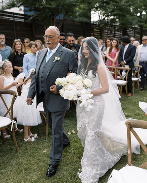 Bride walking down the aisle with her father during an outdoor ceremony. She wears the sheer AMELIE Bridal Veil with blusher over her face, and a fitted lace gown.