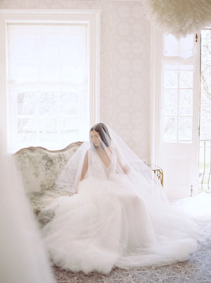 Bride seated on an antique velvet sofa at Graydon Hall Manor. The cathedral length AMELIE Veil with Blusher completely covers her and pools around her tulle ballgown, creating a romantic look.
