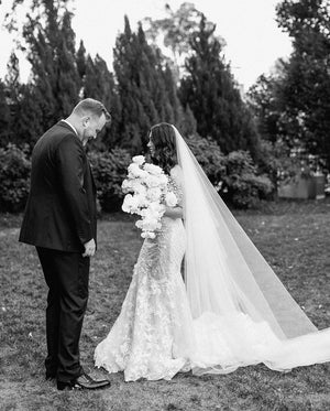 Black and white photo of the bride and groom during their first look outdoors. The bride holds a large bouquet and wears the cathedral-length AMELIE wedding veil over her lace gown.