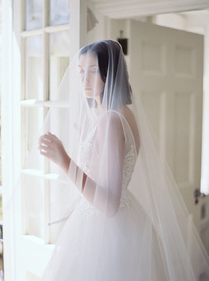 Romantic portrait of a bride standing by a window at Graydon Hall Manor and adjusting the blusher on the AMELIE Bridal Veil.