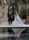 Full-length portrait of a bride wearing the sheer, soft Annabelle English net and a fitted mermaid wedding dress. The bride holds the groom's hand as they stand in an outdoor courtyard, with the long, elegant veil reflecting in the water of a pool in the foreground.