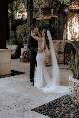 The bride and groom embrace in a semi-covered tropical courtyard. The Annabelle wedding veil flows behind the bride, pooling on the light stone floor.