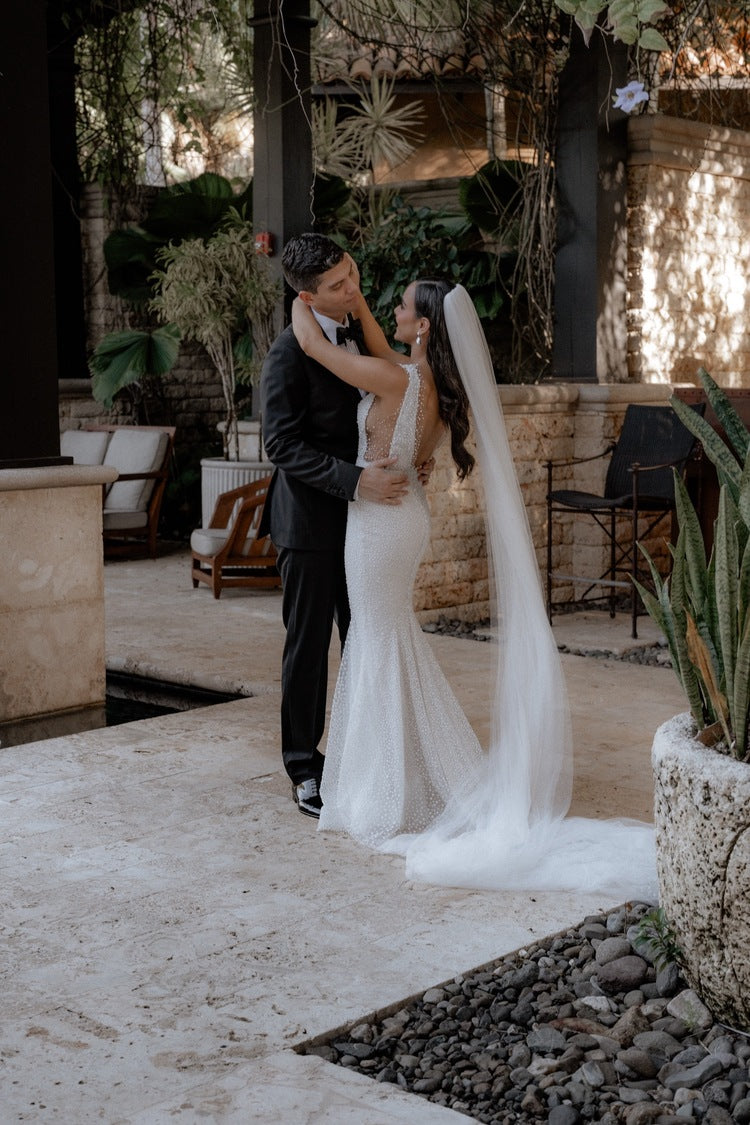 The bride and groom embrace in a semi-covered tropical courtyard. The Annabelle wedding veil flows behind the bride, pooling on the light stone floor.