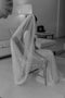 Black and white photo of a bride sitting on a wicker chair indoors. The one-tier Annabelle wedding veil covers her shoulder and back, showcasing the soft drape and sheerness of the luxurious English net. 