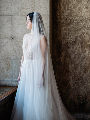 Medium shot of a bride by a window in the Mt Stephen Ballroom at Fairmont Banff. The soft Annabelle English net wedding veil covers her shoulders, demonstrating the veil's sheer quality and softness.
