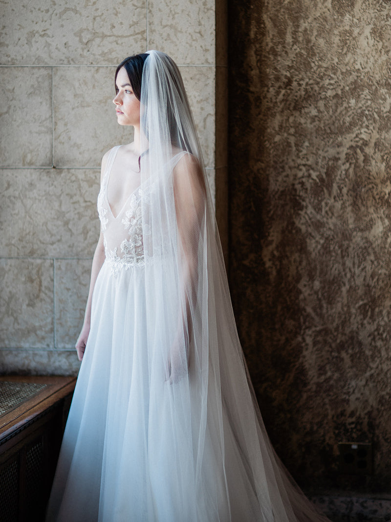 Medium shot of a bride by a window in the Mt Stephen Ballroom at Fairmont Banff. The soft Annabelle English net wedding veil covers her shoulders, demonstrating the veil's sheer quality and softness.