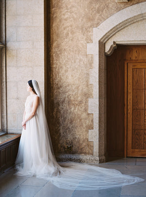 Elegant full-body side view of the bride standing by a tall window in the Mt Stephen Ballroom at Fairmont Banff. The soft Annabelle English net wedding veil drapes down her side, with the extra-long train pooling beautifully on the stone floor.