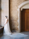 Elegant full-body side view of the bride standing by a tall window in the Mt Stephen Ballroom at Fairmont Banff. The soft Annabelle English net wedding veil drapes down her side, with the extra-long train pooling beautifully on the stone floor.