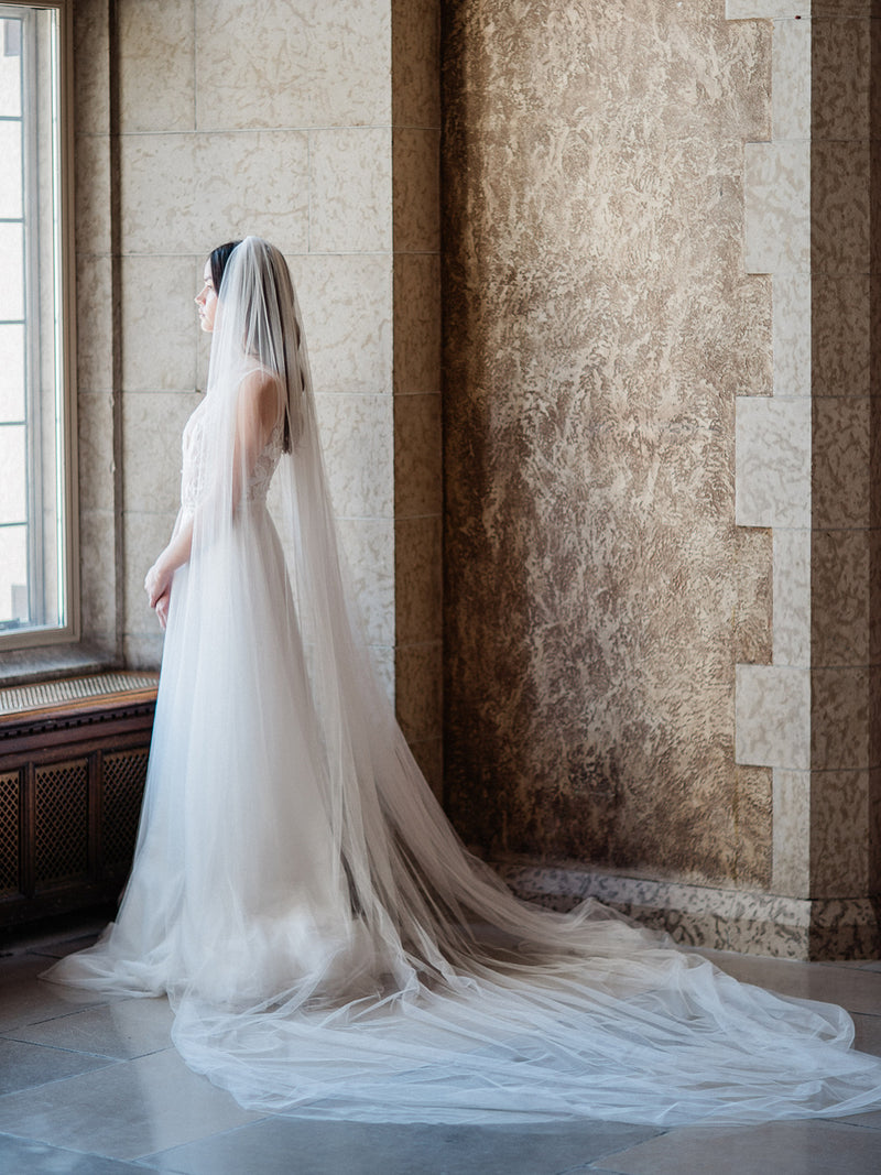 A full height portrait of the bride as she gazes out a window in the Mt Stephen Ballroom at Fairmont Banff. The single-tier Annabelle English net wedding veil is spread out behind her to show the  dramatic width of the train.