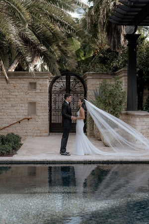 Bride and groom standing facing each other next to a pool. The long, elegant Annabelle English Net wedding veil softly flows behind the bride.