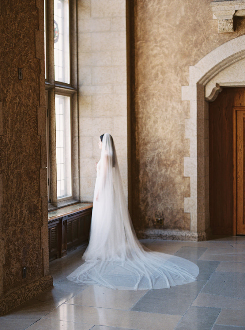 Wide shot showing the full length of the Annabelle English net wedding veil in the Mt Stephen Ballroom at Fairmont Banff. The veil's sheer train is widely fanned out on the floor behind the bride as she stands looking out the window.