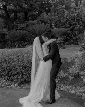 A romantic black and white photo of the bride and groom embracing outdoors. The sheer, soft Annabelle wedding veil drapes delicately down the bride's back, emphasizing the deep V-back of her dress.