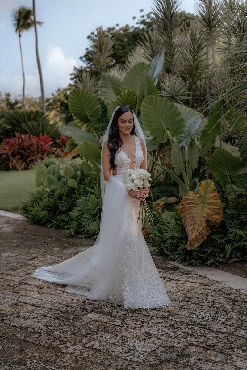 A full-body shot of the bride walking through a lush, tropical garden. The Annabelle wedding veil trails behind her on the stone path. The veil is placed on the crown of the head, highlighting the sleek, side-parted hair down hairstyle. 