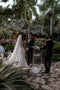 The wedding ceremony in a lush, outdoor palm garden. The bride, viewed from the back, wears the long, single-tier Annabelle English net wedding veil, which flows down dramatically.