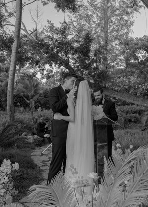 Black and white photo of the bride and groom kissing at the altar during their outdoor ceremony, while the officiant stands nearby. The Annabelle English net wedding veil softly drapes down the bride's back.