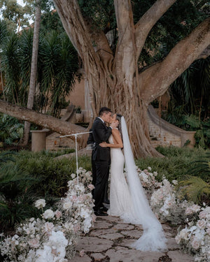 The bride and groom share a kiss under a large, striking tree, surrounded by floral arrangements. The Annabelle cathedral wedding veil flows down the bride's back, blending with the soft floral decor at the ceremony site.
