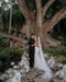 The bride and groom share a kiss under a large, striking tree, surrounded by floral arrangements. The Annabelle cathedral wedding veil flows down the bride's back, blending with the soft floral decor at the ceremony site.