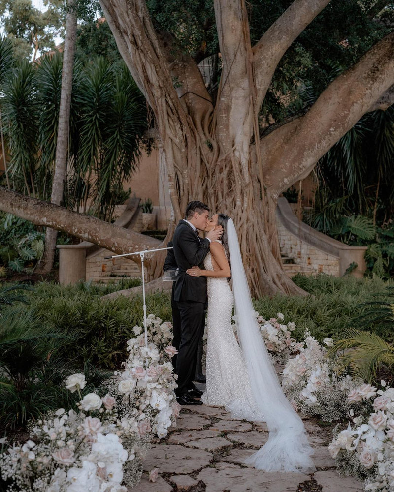 The bride and groom share a kiss under a large, striking tree, surrounded by floral arrangements. The Annabelle cathedral wedding veil flows down the bride's back, blending with the soft floral decor at the ceremony site.