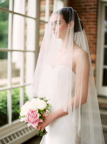 Bride wearing the ARIA sheer wedding veil with blusher; she's wearing a strapless crepe wedding dress, has her hair in an updo, and is holding an ivory and blush peony bouquet.