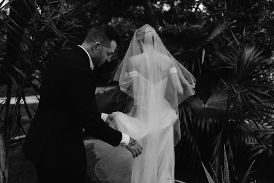 Bride wearing the ARIA sheer wedding veil with blusher with an off-the-shoulder wedding dress. Her groom is helping carry the cathedral length veil train. 