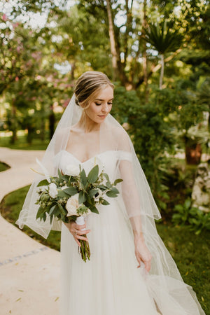 Bride wearing the ARIA sheer wedding veil with blusher with an off-the-shoulder wedding dress; her hair is styled in a soft updo and she's holding a white and green bouquet.