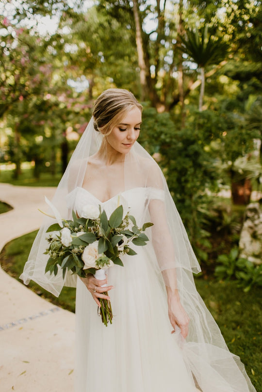 Bride wearing the ARIA sheer wedding veil with blusher with an off-the-shoulder wedding dress; her hair is styled in a soft updo and she's holding a white and green bouquet.