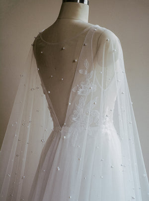 Close-up, rear view of the ARIELLE pearl bridal cape draped over a mannequin wearing a wedding gown. The sheer tulle is scattered with pearls, highlighting the deep V-back of the dress underneath.