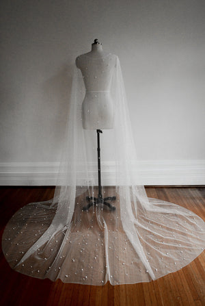 Full-length view of the ARIELLE pearl bridal cape draped on a mannequin, with the chapel train spread out on the wooden floor, illustrating the scale and sheer effect.