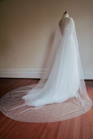 Back view focusing on the extensive, pearl-scattered chapel train of the ARIELLE bridal cape as it pools elegantly on the floor.