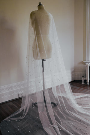 Three-quarter view of the ARIELLE cape on a mannequin, showing the full length and the luxurious sweep of the pearl-embellished tulle pooling on the hardwood floor.