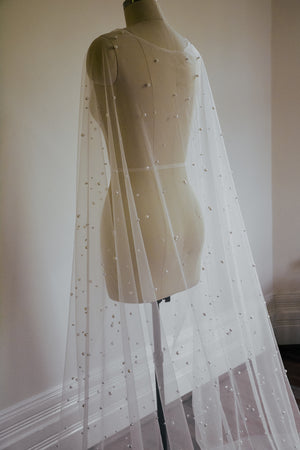 Close-up of the ARIELLE pearl cape hanging on a mannequin, focusing on the sheer fabric and the density of the pearl scattering down the length of the cape.